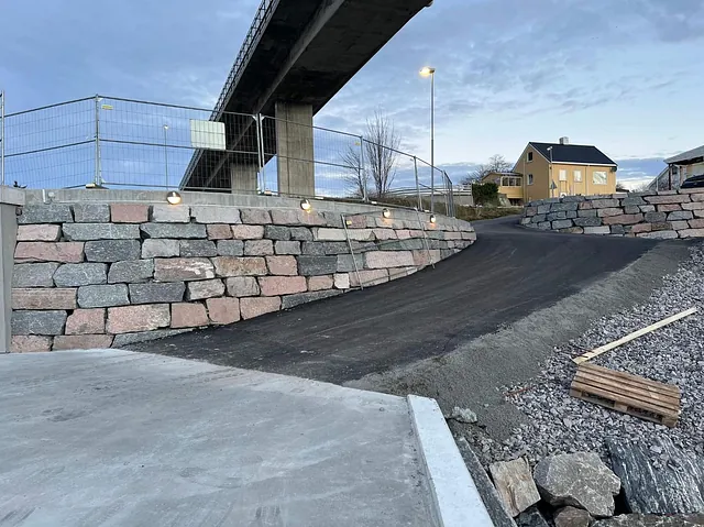 Nyasfaltert rampe med solide natursteinsmurer under bro, belysning montert i betongkanten.