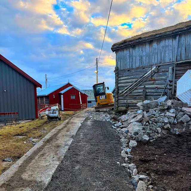 Grunnarbeid pågår ved en gammel laftet bygning hvor steinmasser fjernes, og ny gangvei etableres med minigraver på stedet.