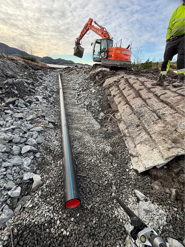 Nærbilde av grøft med sort plastledning og grus, gravemaskin i arbeid og person i arbeidsklær til høyre.