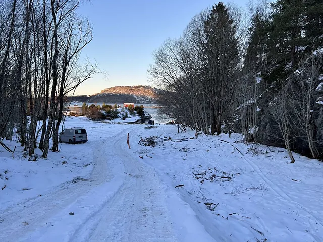 Snødekt landskap med traktorveier som leder ned mot vannet, omgitt av trær og fjell i bakgrunnen.