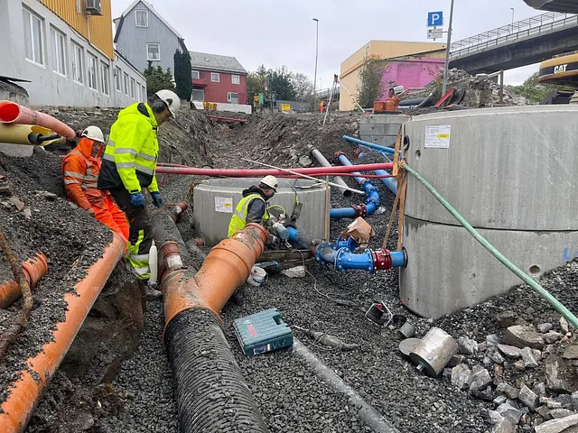 Anleggsarbeidere monterer vann- og avløpsrør til nye kummer i tettbebygd område, med omfattende rørinstallasjoner og koordinering pågår.