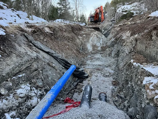 Grøft med nedlagte rør i ulike størrelser og gravemaskin i bakgrunnen, omgitt av snødekt terreng.