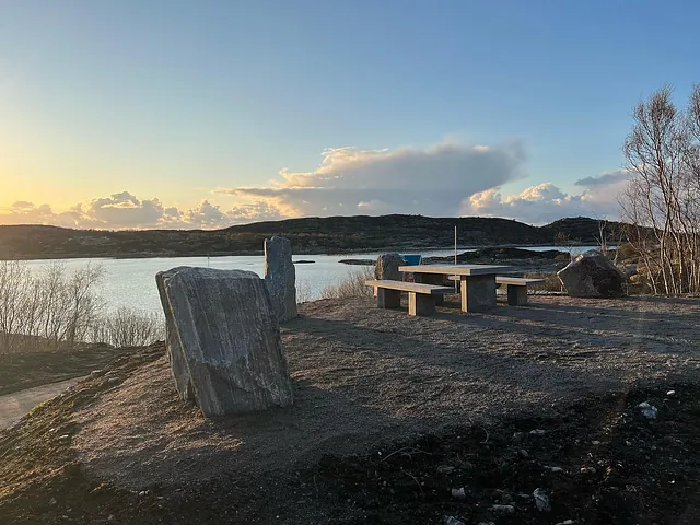 Uteplass med steinbord og benker ved sjøen, omgitt av svaberg og kveldssol i horisonten.