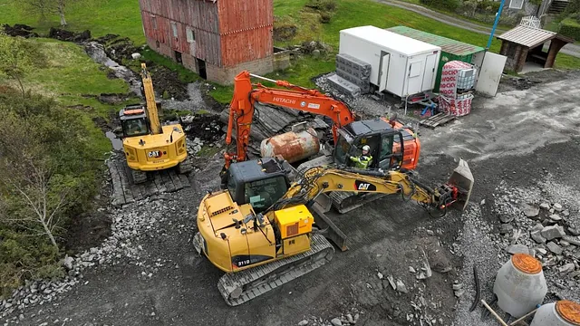 Fire gravemaskiner jobber tett ved et gammelt bygg og riggområde med containere og byggematerialer.