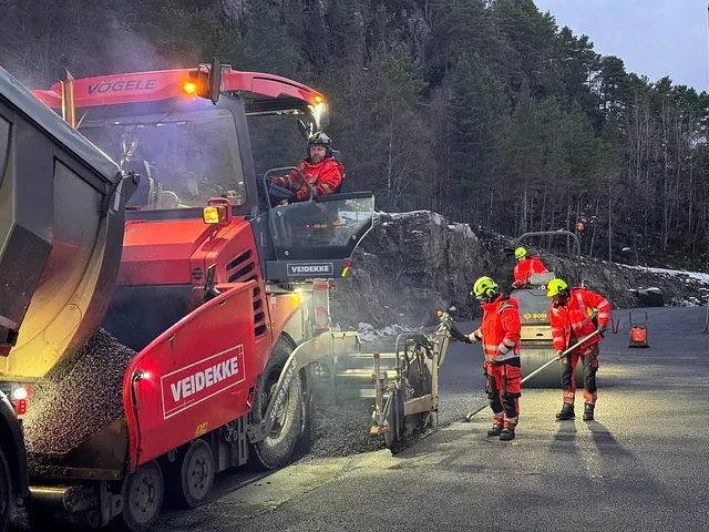 Asfalteringsarbeid med asfaltutlegger og mannskaper i arbeid ved fjellside i vinterlige omgivelser.