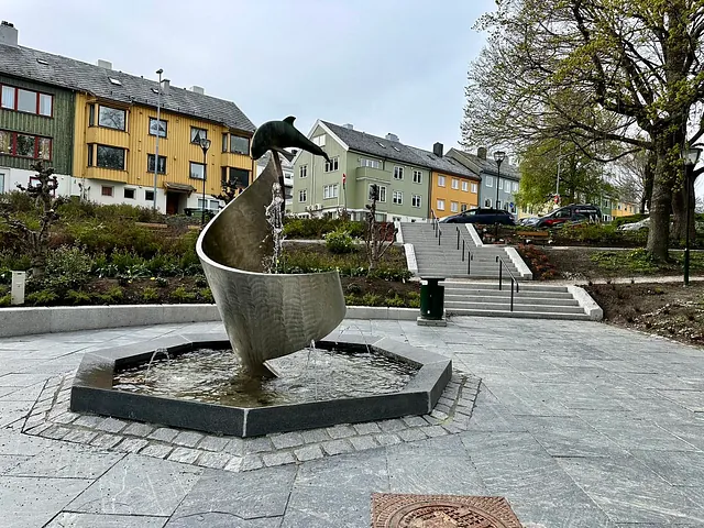En offentlig park med fontene formet som en bølge med en delfin på toppen. Vann renner ned i et åttekantet basseng. I bakgrunnen ses fargerike trehus og en trapp som leder opp til et grøntområde med trær og parkerte biler.