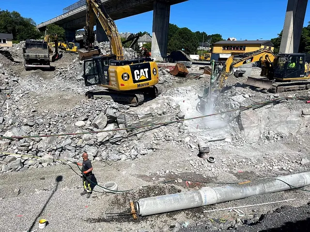 Større anleggsområde med gravemaskiner og riving av fjell under bro i tettbebygd strøk.