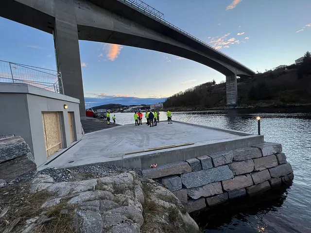 Gruppe med arbeidere ved ny brygge under bro, med vann og by i bakgrunnen.