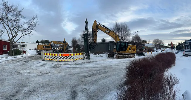 Gravemaskiner i arbeid med grunnboring og graving i et boligområde med snødekke og vinterstemning.