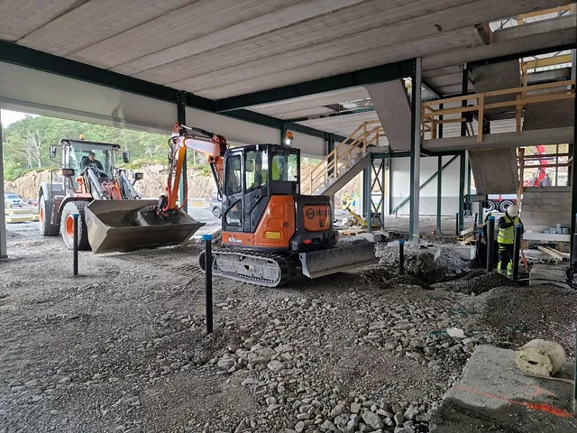 Grunnarbeid inne i industribygg under oppføring, med maskiner og arbeidere i aktivitet.
