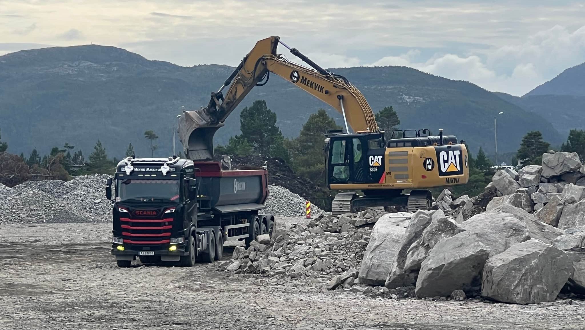 Gul CAT gravemaskin med Mekvik logo løfter stein og grus over i lastebil