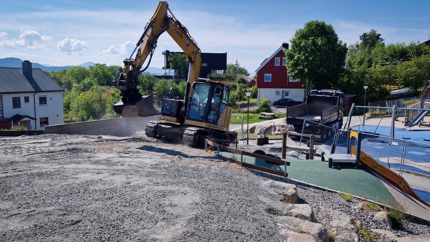 Gravemaskin arbeider med utgraving nær et inngjerdet lekeområde i et boligområde med hus og trær i bakgrunnen.