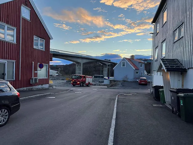 Gatebilde med nylagt asfalt, bolighus og brannbil nær byggeplass ved bru i bakgrunnen.