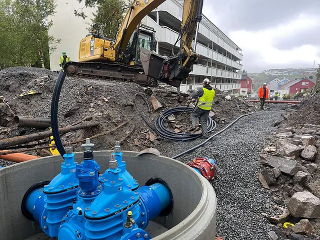 Gravemaskin og arbeidere legger trekkerør og vannrør ved leilighetsbygg. Ny vannkum med ventiler installert i forkant.