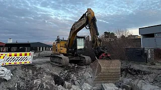 Gravemaskin graver i steinete grunn nær boligområde, omgitt av sperringer og vinterkledd landskap.