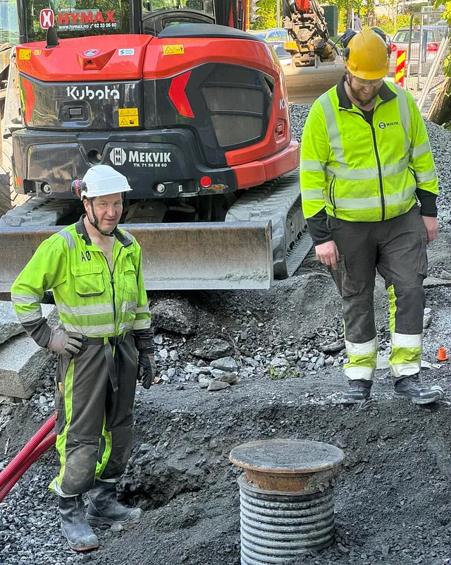 To arbeidere på anleggsområde iført atbeidsklær står foran rød gravemaskin