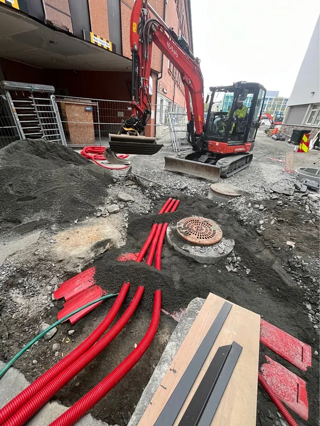 Minigraver og elektriske rør klargjøres i gategrunn ved bygg med rødfasade.