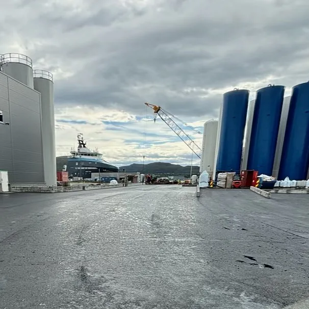 Asfaltert industriområde ved kai med store siloer og supplyskip i bakgrunnen under overskyet himmel.