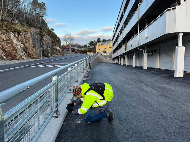 Arbeider i refleksjakke inspiserer rekkverk ved nyasfaltert vei foran boligblokker.