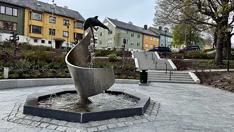 En offentlig park med fontene formet som en bølge med en delfin på toppen. Vann renner ned i et åttekantet basseng. I bakgrunnen ses fargerike trehus og en trapp som leder opp til et grøntområde med trær og parkerte biler.