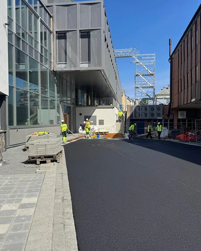 Asfaltering i byrom ved nytt bygg, med arbeidere i aktivitet og stillas i bakgrunnen.