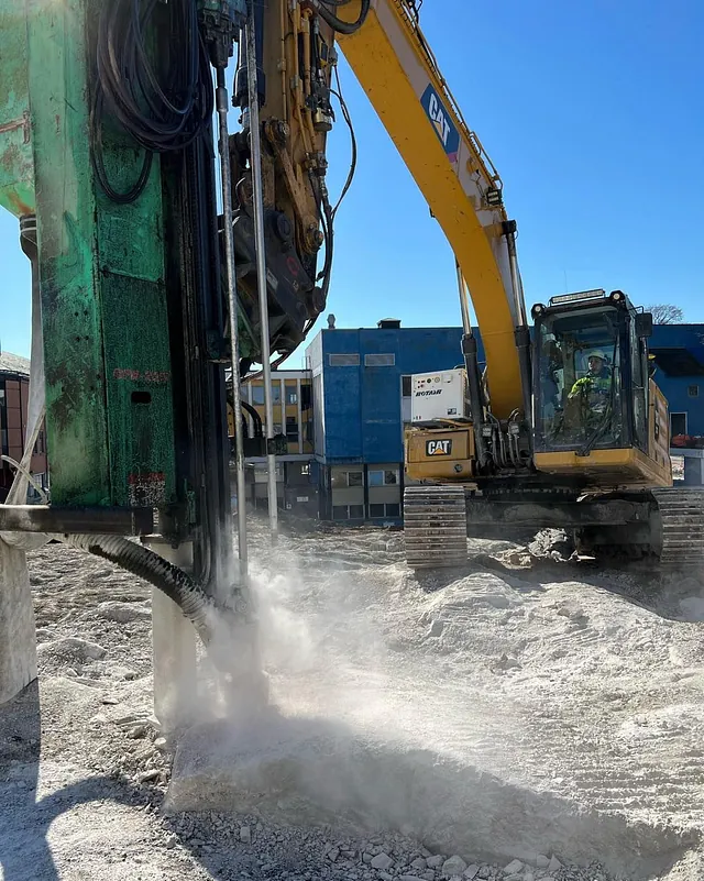 Fundamenteringsarbeid med borerigg i gang på fjellgrunn, støv og kraftig maskineri i aksjon.
