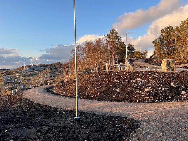 Svingete gangvei i kystterreng med gatelykter, trær og steinblokker langs stien.