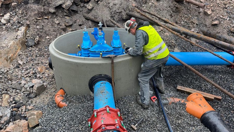 Anleggsarbeider jobber med montering av store blå vannrør og ventiler i en kum i gruslagt grøft.
