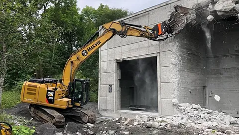 Gravemaskin fra Mekvik river en betongvegg på en bygning, med støv og stein som faller ned i forgrunnen.