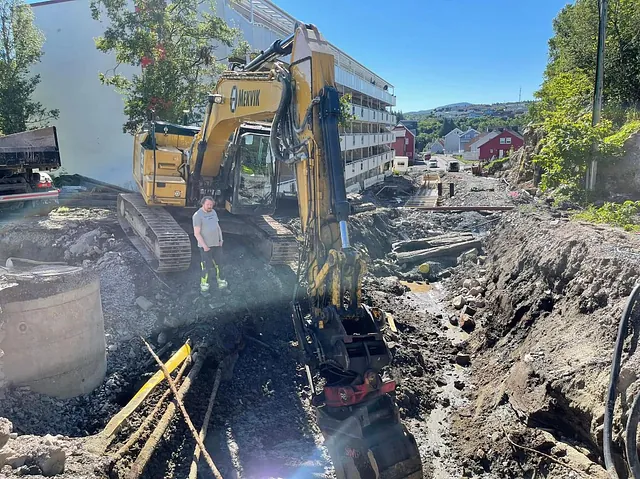 Mann ved stor gravemaskin i grøft fylt med rør, stein og vann nær boligblokker.