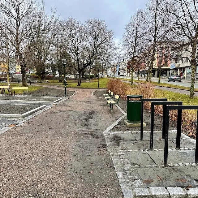 Gruslagt parksti med benker, søppelbøtter og nakne trær langs en gate med butikker.
