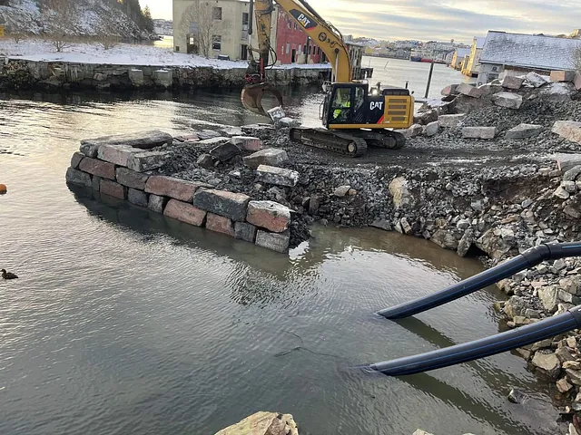 Steinfylling og gravemaskin i arbeid med å bygge molo eller kaifront i vannkanten.