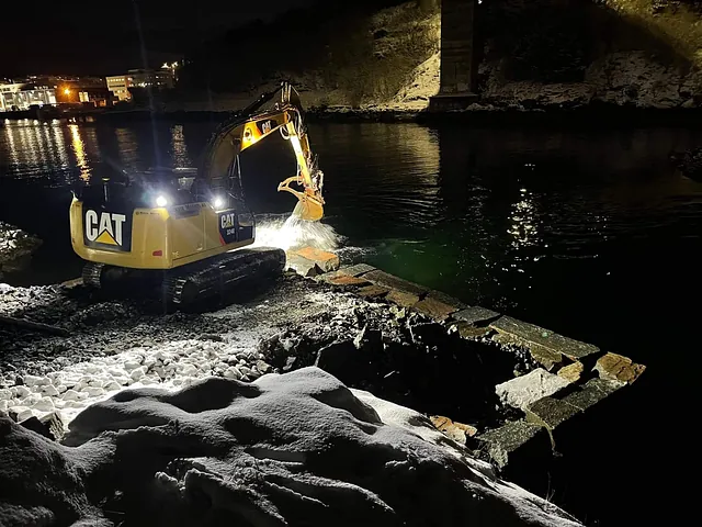 Gravemaskin legger stein i vannkanten i mørket, opplyst av maskinens frontlys.