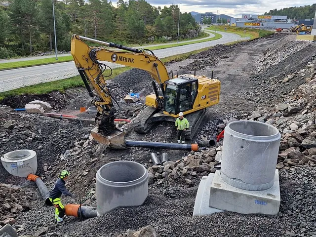 Gravemaskin og anleggsarbeidere installerer store betongrør ved vei i skrånende terreng.