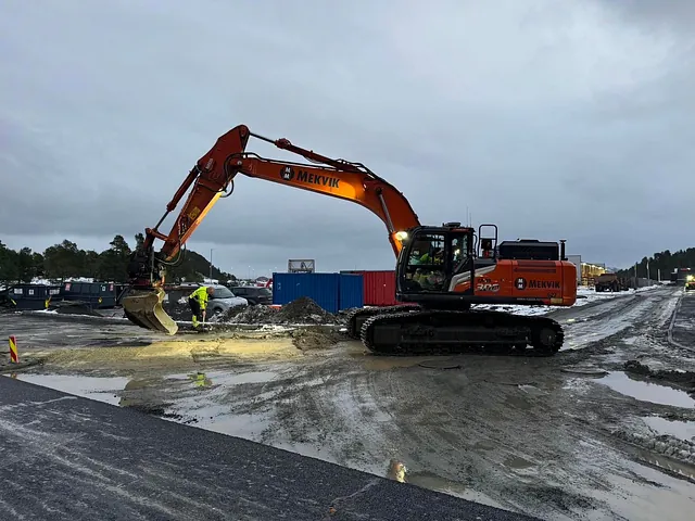 Gravemaskin og anleggsarbeidere i arbeid med grunnarbeid på en våt og sølete anleggsplass.