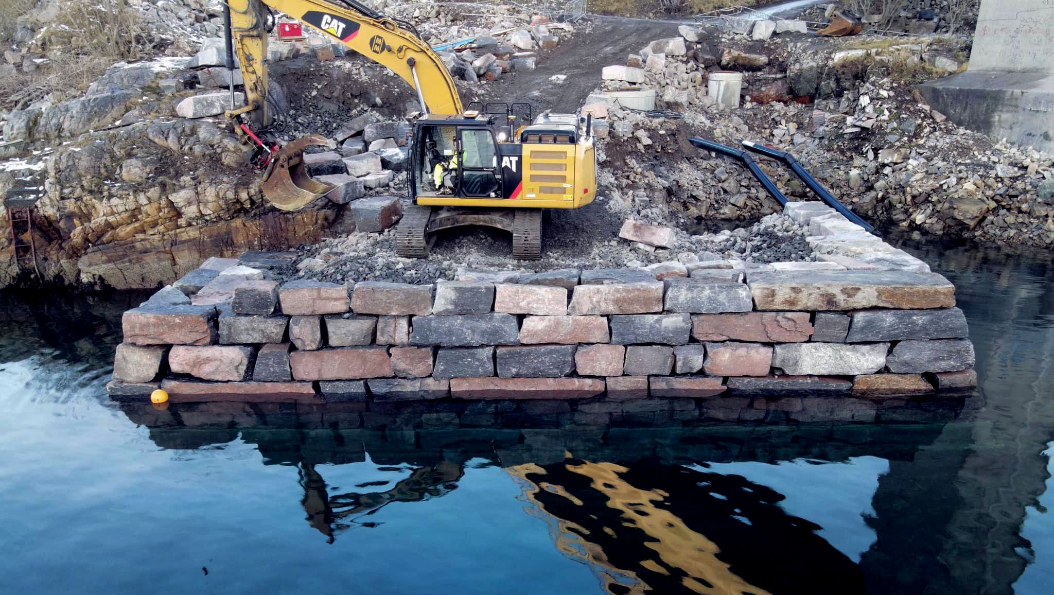 Gravemaskin står på platå bygget ut i sjøen med steinmur rundt.
