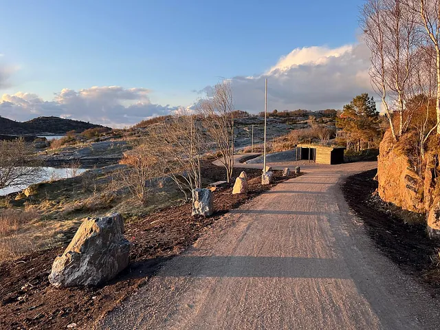 Gruslagt turvei langs vannet med steinblokker og gapahuk, omgitt av lav vegetasjon.