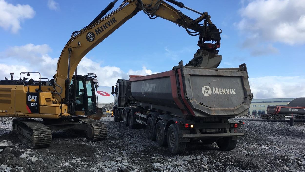 Gravemaskin fra Mekvik laster stein i en lastebil på anleggsplass med grus og maskiner i bakgrunnen.