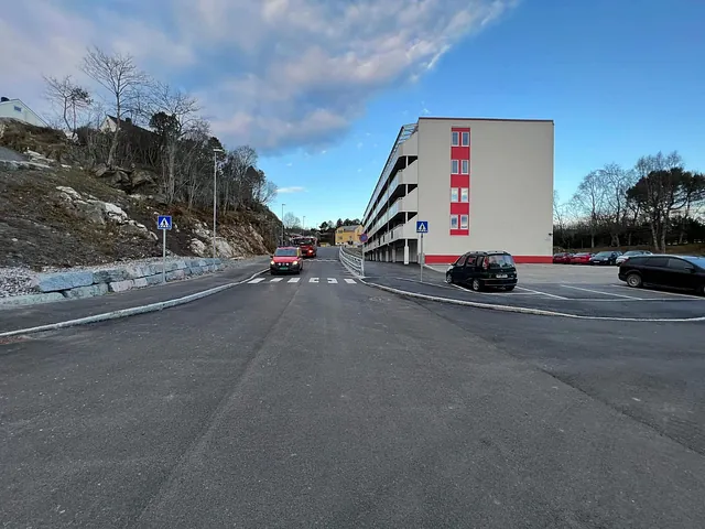 Nyasfaltert vei med fortau, fotgjengerfelt og boligblokk på høyre side, biler i trafikken.