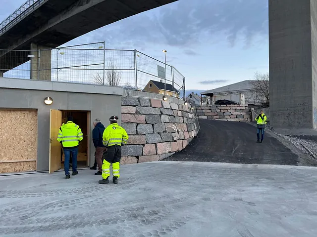 Arbeidere og byggeleder inspiserer nytt teknisk bygg under brua. Adkomst er etablert med støttemur og asfaltert vei.