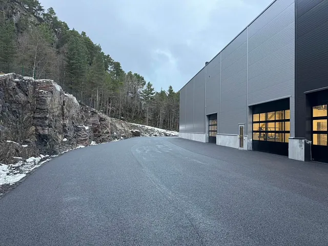 Nyasfaltert vei ved en stor industribygning, med fjellskjæring og skog i bakgrunnen.