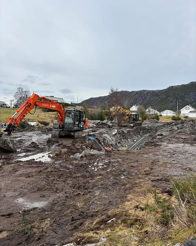 To gravemaskiner arbeider i et gjørmete terreng med grøft og ledninger, omgitt av fjell og hus.