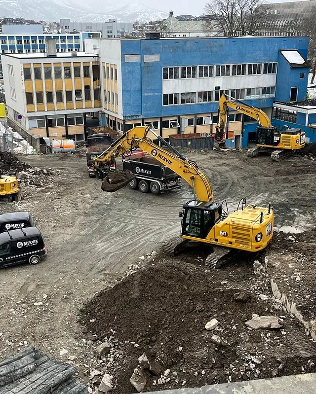 Grunnarbeid med gravemaskiner og lastebil foran eldre bygg, snødekte fjell i bakgrunnen.