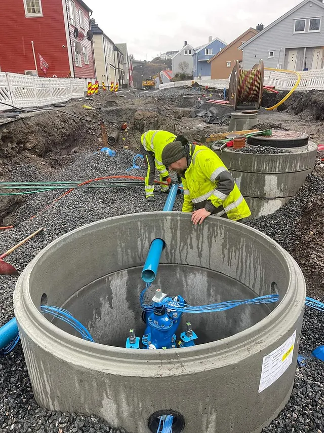 To arbeidere monterer blå vannrør i betongkummer under anleggsarbeid i en boliggate.