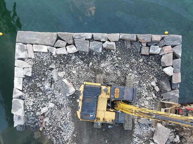 Gravemaskin sett ovenfra på molo under oppbygging med store steinblokker i klart vann.